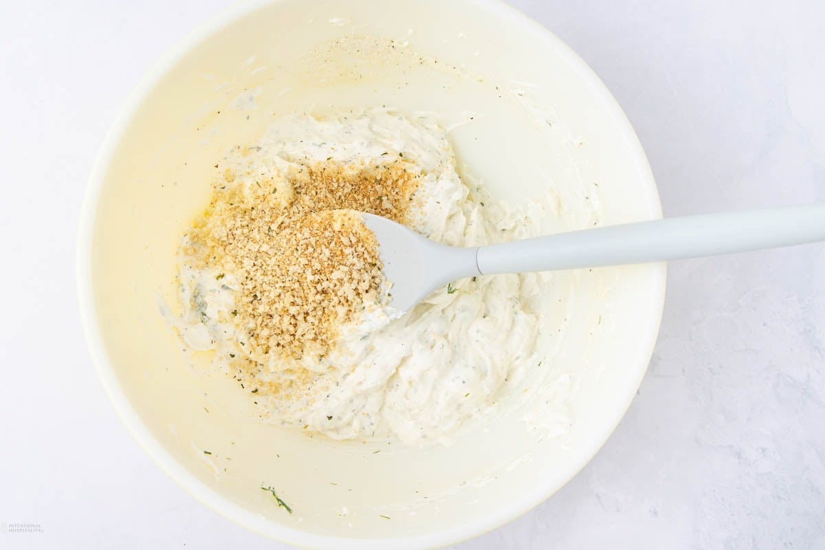 A mixing bowl containing creamy white dip with a sprinkle of brown seasoning on top. A spatula rests in the bowl, partially covered by the dip. The bowl is set on a light, textured surface.