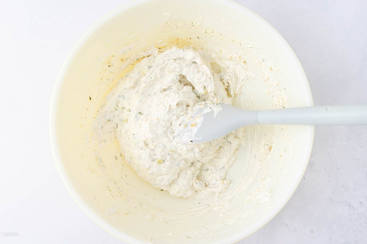 A white mixing bowl containing a creamy mixture with herbs, being stirred with a gray spatula. The consistency appears smooth and seasoned, possibly a dip or spread. The bowl sits on a light-colored surface.