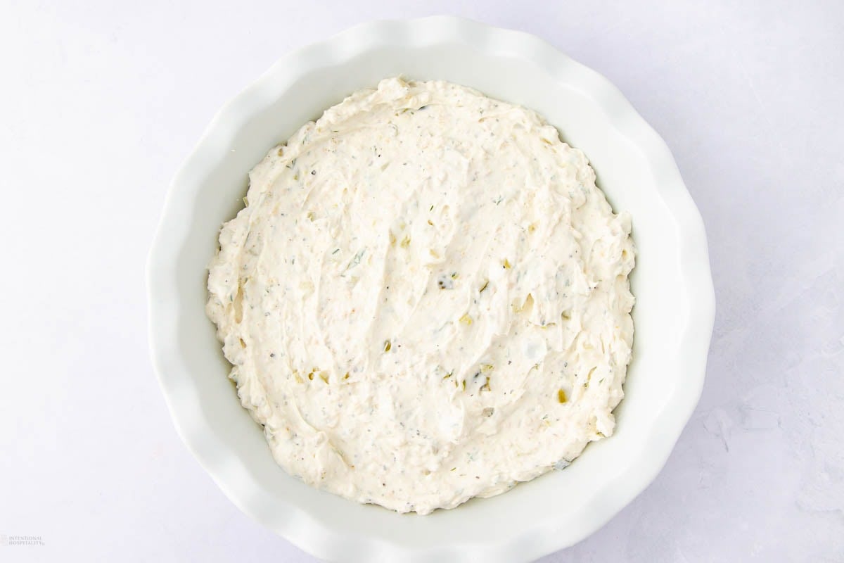 A white pie dish filled with a creamy cheese mixture, flecked with herbs, on a light gray surface.