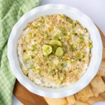 A round white dish filled with a creamy dip topped with chopped dill, breadcrumbs, and sliced pickles. It's placed on a wooden board with pita chips beside it. A green checkered cloth is partially visible in the corner.