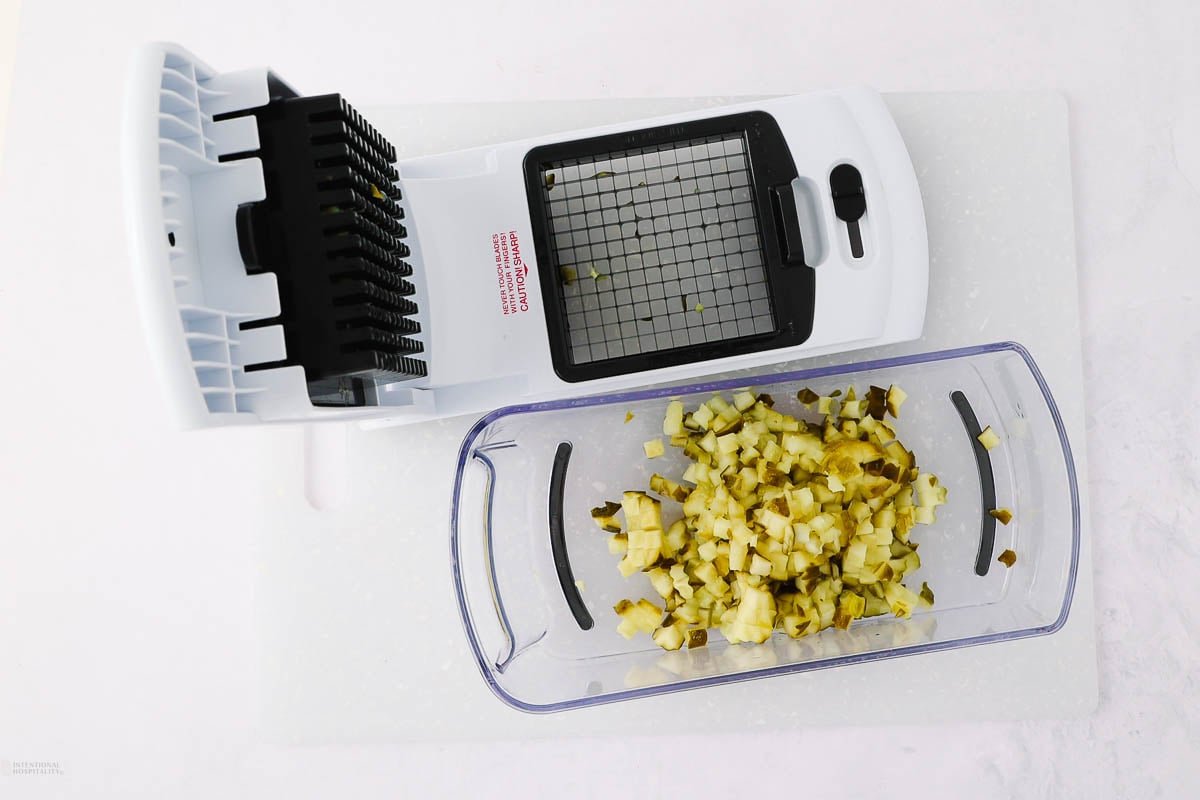 A white vegetable chopper with a grid blade is placed on a white surface. Diced yellow vegetables, possibly pickles, are collected in a transparent container at the base of the chopper.