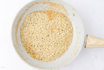 A frying pan with toasted breadcrumbs. The breadcrumbs are golden brown and evenly spread across the pan. The pan has a light-colored, speckled non-stick surface with a wooden handle, set on a white background.