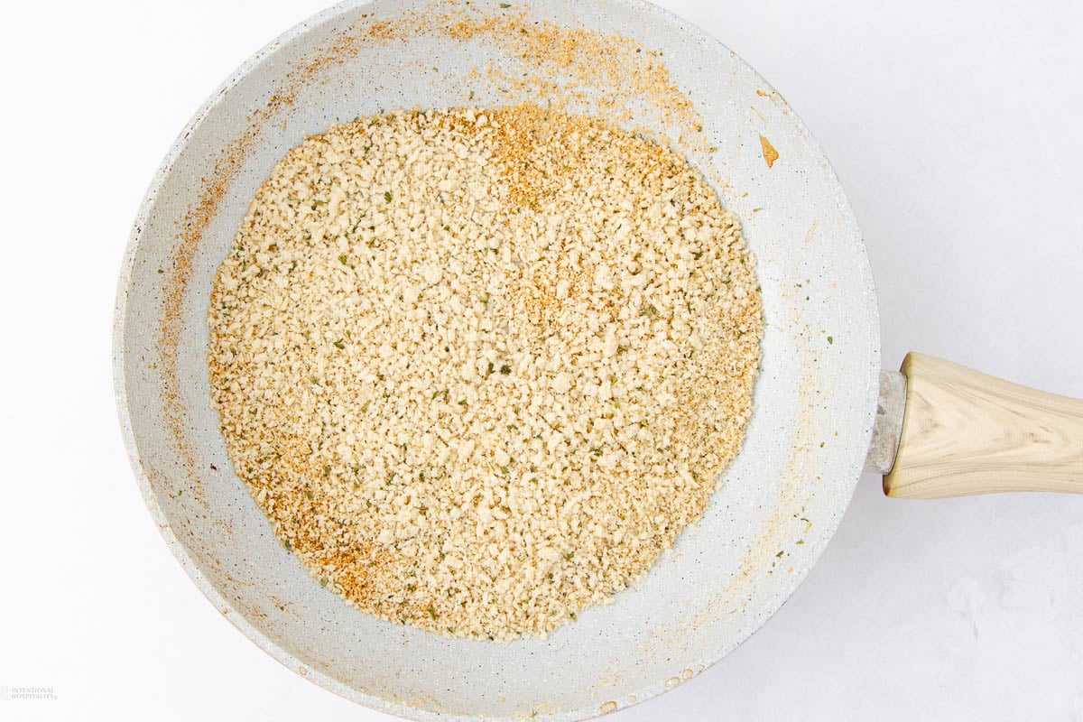 A frying pan with toasted breadcrumbs. The breadcrumbs are golden brown and evenly spread across the pan. The pan has a light-colored, speckled non-stick surface with a wooden handle, set on a white background.