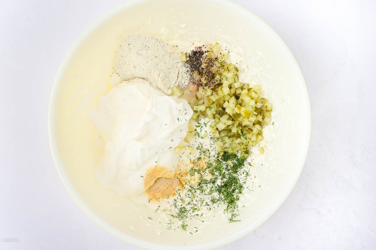 A mixing bowl containing yogurt, diced pickles, dry spices, herbs, and a seasoning blend. The ingredients are ready to be mixed together, likely for a dip or sauce. The background is a plain, light surface.