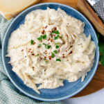 A blue ceramic plate filled with creamy fettuccine Alfredo, garnished with chopped parsley and red pepper flakes. A block of Parmesan cheese and a grater are nearby on a wooden surface. A textured cloth napkin in the background.