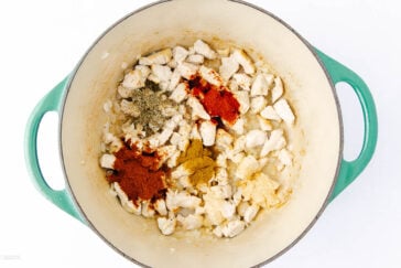 A large green pot filled with chopped chicken pieces and various spices, including chili powder, cumin, garlic powder, and dried herbs, ready to be mixed. The pot is on a white background.