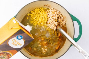 A pot of soup being prepared with corn and white beans visible. Chicken broth is being poured from a box into the pot, which also contains pieces of chicken and herbs. A spoon is in the pot, and the pot is green with a worn handle.