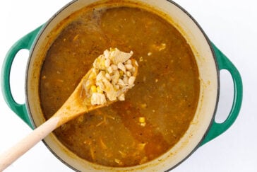 A green pot filled with a chunky soup, including pieces of chicken, white beans, corn, and a rich broth. A wooden spoon lifts a portion of the soup, showcasing its ingredients.