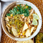 A ceramic bowl filled with chili topped with tortilla chips, sliced jalapeños, fresh cilantro, and lime wedges. The chili contains white beans, corn, and other ingredients, all resting on a woven placemat.