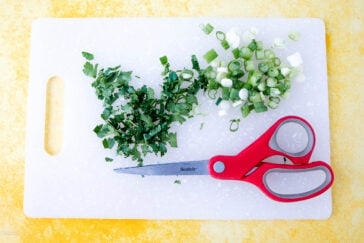 Chopped cilantro and green onions rest on a white cutting board, ideal for preparing a canned cowboy candy recipe. Red-handled scissors lie nearby, ready for action. The backdrop is a textured yellow surface, adding vibrant contrast to this culinary scene.