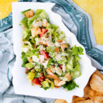 A salad with chopped lettuce, tomatoes, croutons, and shaved parmesan is served on a Georgia-shaped ceramic plate. It rests on a blue and white cloth, with canned cowboy candy nearby for added zest and some pita chips within reach.