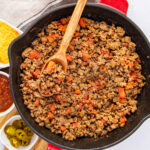 A skillet filled with cooked ground beef and diced tomatoes, topped with a wooden spoon. In the background, there are small dishes containing shredded cheese, salsa, and sliced jalapeños.