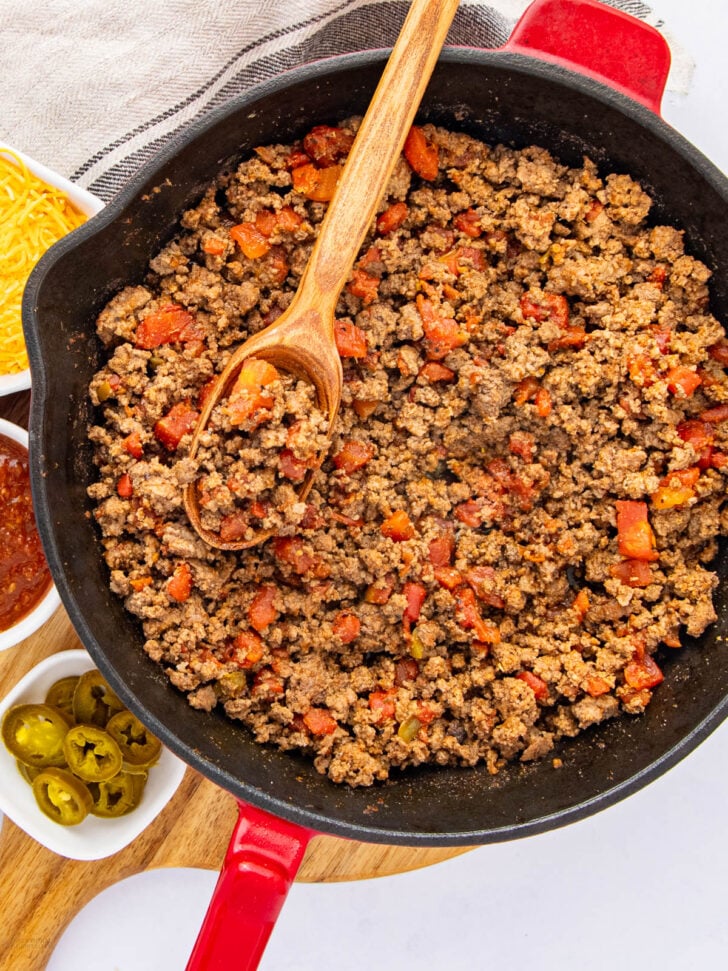 A skillet filled with seasoned ground beef and diced tomatoes, with a wooden spoon resting on top. Surrounding the skillet are small bowls containing shredded cheese, salsa, and sliced jalapeños.