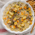 A white dish filled with a creamy stew featuring diced potatoes, carrots, ground meat, peas, and green beans. The stew is served next to grilled bread slices, all placed on a cloth with red stripes.
