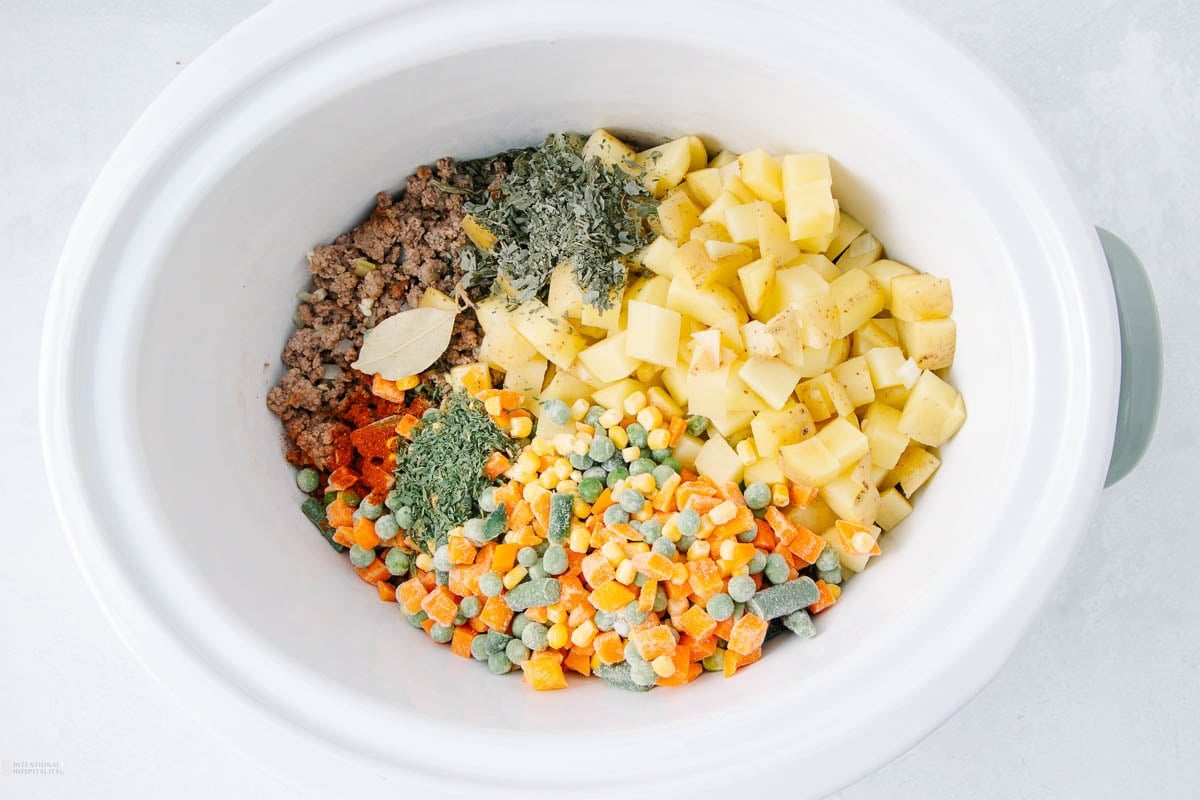 A slow cooker filled with uncooked ground meat, diced potatoes, mixed frozen vegetables, herbs, and a bay leaf, all arranged in separate sections on the bottom of the white pot.