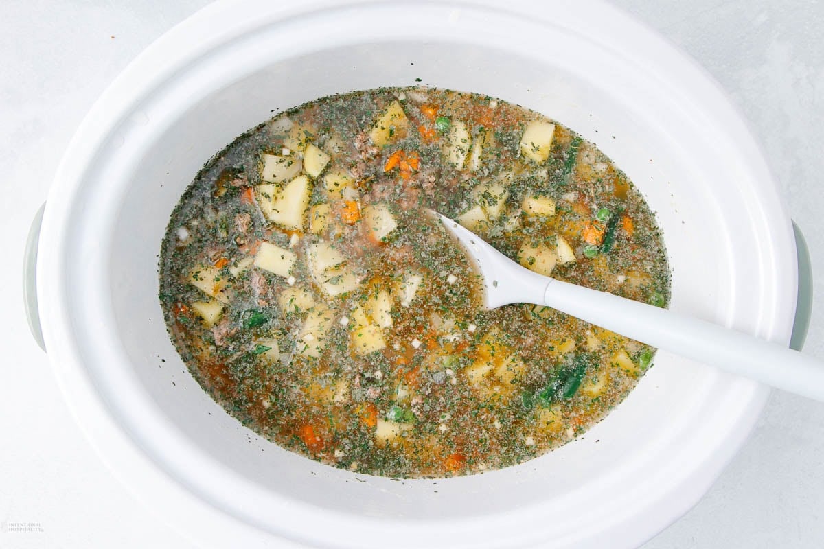 A white slow cooker filled with a hearty soup, including visible chunks of potatoes, carrots, and herbs in a broth. A white ladle is resting inside the pot, ready to serve.