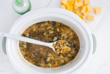 A white slow cooker filled with vegetable soup, containing diced carrots, potatoes, and herbs. A white ladle scoops some of the soup. Cubes of cheese are placed on a cutting board nearby.
