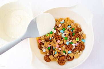 A mixing bowl filled with a variety of snacks, including pretzels, colorful candy-coated chocolates, cereal pieces, and peanuts. A spatula coated with white melted chocolate rests on the bowl's edge.
