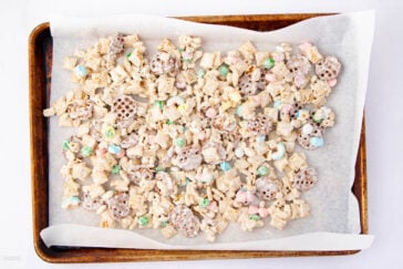 A baking sheet lined with parchment paper is filled with a mixture of cereal, pretzels, and colorful candy-coated pieces, all coated in a white glaze.