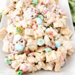 A close-up of a snack mix on a white plate, featuring cereal squares, pretzels, colorful candy-coated chocolates, and marshmallows coated in a white, sweet glaze. The texture appears crunchy and sweet.