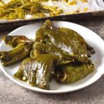 A white plate brimming with roasted green peppers glistens with a hint of oil and is garnished with seeds, reminiscent of a savory twist on canned cowboy candy. In the background, a tray holding more roasted peppers can be seen on the textured gray surface.