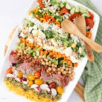 A large platter of colorful chopped salad with rows of ingredients: cucumbers, peppers, cheese, eggs, nuts, chickpeas, meats, radishes, tomatoes, seeds, and greens. Wooden serving utensils and a green cloth are beside the platter.