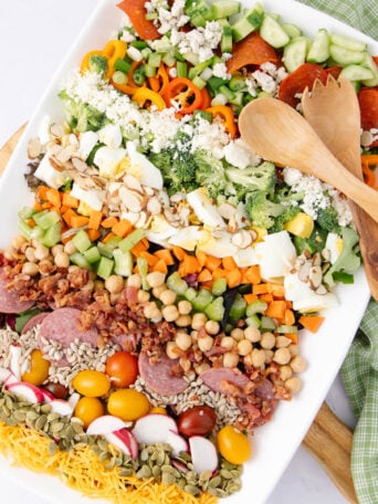 A large platter of colorful chopped salad with rows of ingredients: cucumbers, peppers, cheese, eggs, nuts, chickpeas, meats, radishes, tomatoes, seeds, and greens. Wooden serving utensils and a green cloth are beside the platter.