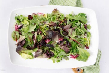 A rectangular white plate filled with a variety of fresh, colorful mixed salad greens. The greens rest on a green checked napkin on a light surface.