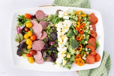 A colorful salad on a rectangular white plate with mixed greens, sliced salami, cherry tomatoes, chopped eggs, broccoli, bell peppers, pepperoni, and green onions. A green checkered cloth is partially visible underneath the plate.