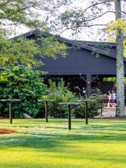 A black barn-like building surrounded by lush green trees and bushes, with people gathered under a covered patio enjoying the masters food, as sunlight illuminates the grassy lawn in the foreground.
