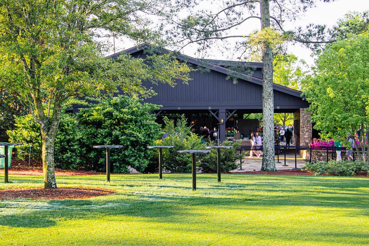 A black barn-like building surrounded by lush green trees and bushes, with people gathered under a covered patio enjoying the masters food, as sunlight illuminates the grassy lawn in the foreground.