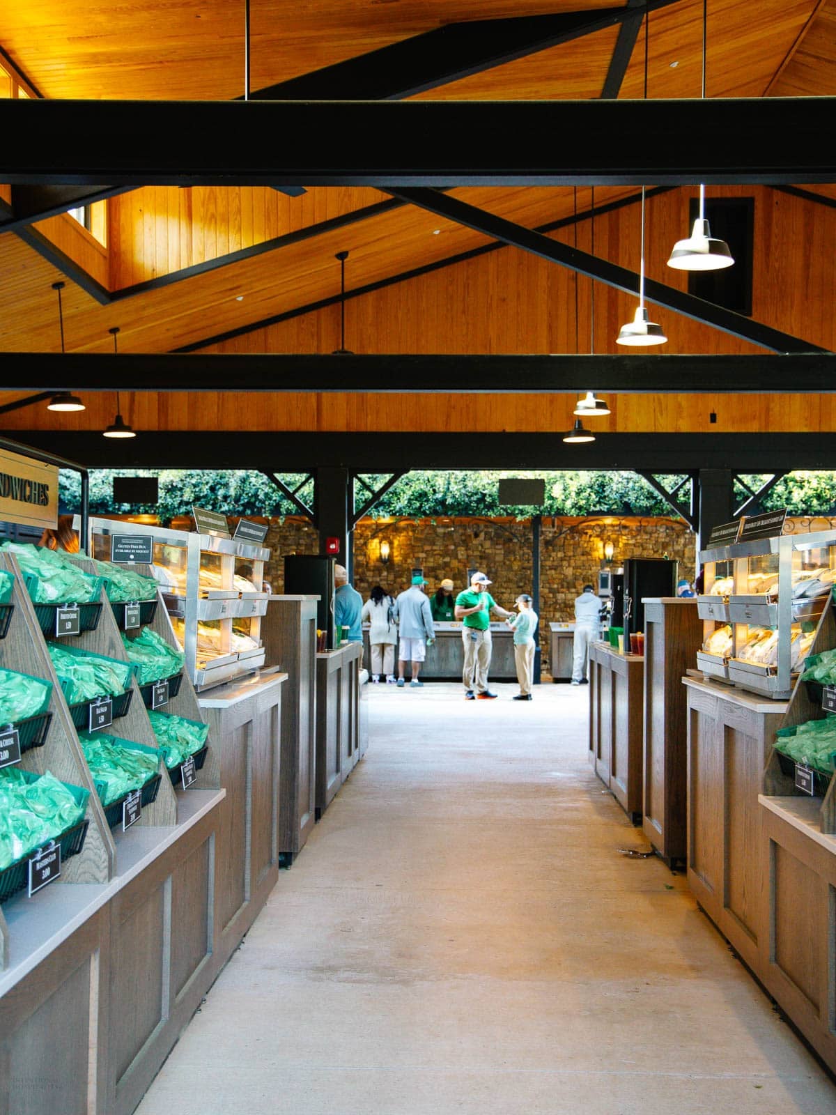 A spacious, well-lit bakery with wooden beams, shelves of bread loaves on both sides, and a group of people ordering at a counter in the background. Natural light and greenery enhance the cozy atmosphere, inviting you to enjoy the masters food.