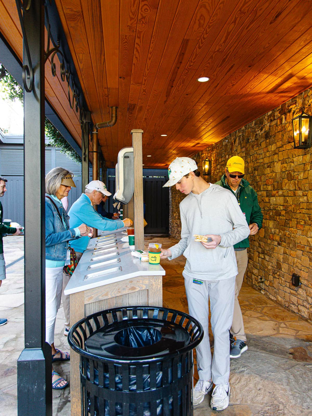 People stand at a row of water fountains along a stone wall under a wooden ceiling. A trash can is in the foreground, and some individuals hold cups and snacks from the Masters food while filling their drinks.