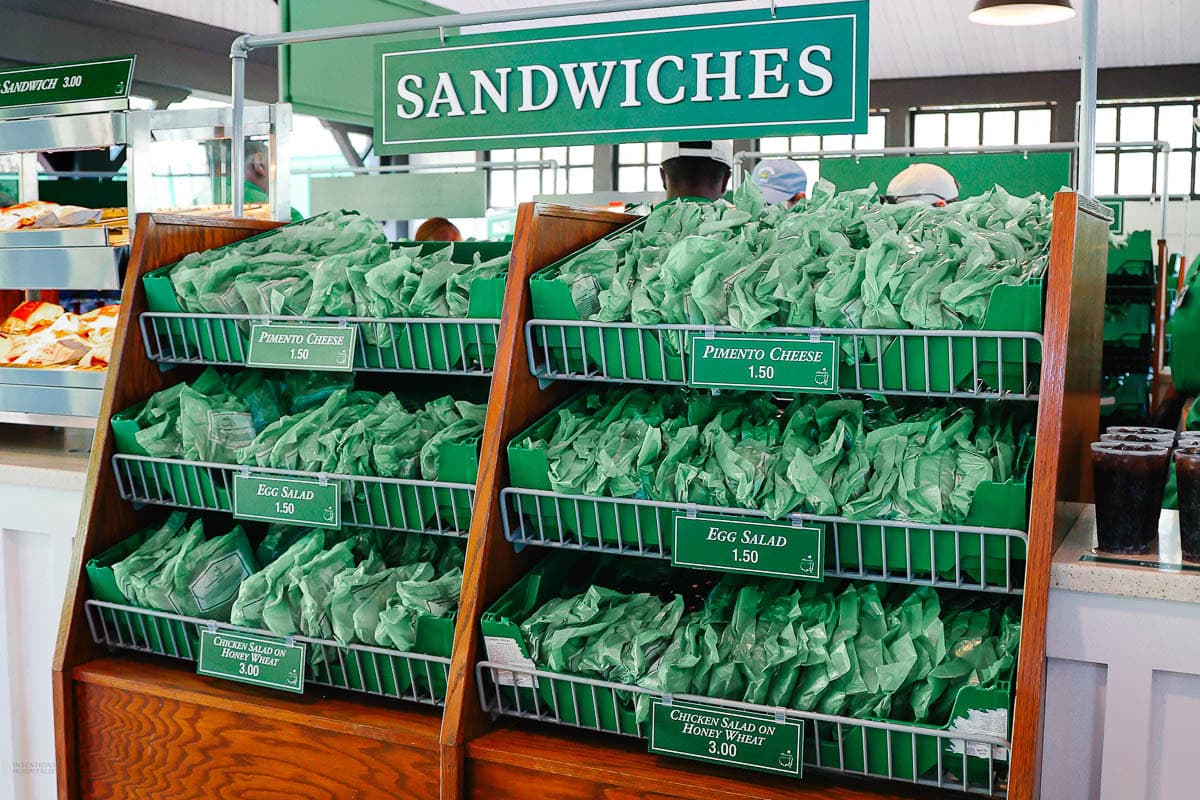 A sandwich display with green-wrapped sandwiches labeled pimento cheese, egg salad, and chicken salad on honey wheat bread, evoking classic the masters food, arranged neatly on wooden shelves under a large green “Sandwiches” sign.
