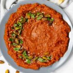 A plate of red tomato dip garnished with green herbs, whole pistachios, and sesame seeds, set against a light background. A napkin is partially visible on the left side.