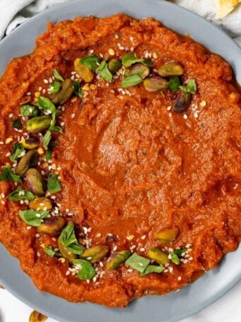 A plate of red tomato dip garnished with green herbs, whole pistachios, and sesame seeds, set against a light background. A napkin is partially visible on the left side.
