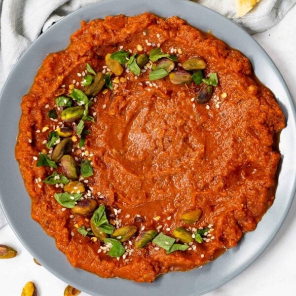 A plate of red tomato dip garnished with green herbs, whole pistachios, and sesame seeds, set against a light background. A napkin is partially visible on the left side.
