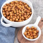 Two bowls of roasted chickpeas seasoned with spices. The larger bowl is white and placed on a blue cloth, while the smaller white spoon-shaped bowl sits on a wooden board. The chickpeas are golden brown.