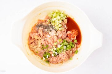 A white bowl contains ground meat mixed with chopped celery, green onions, and various spices, including pepper and garlic powder. A reddish sauce is also present, ready to be mixed. The bowl sits on a white background.