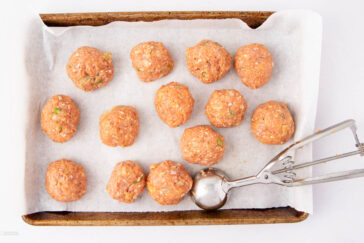 Raw meatballs on a parchment-lined baking tray with a metal scoop on the side.