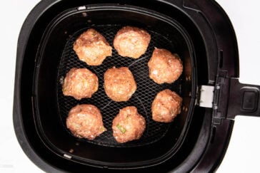 Eight raw meatballs are arranged inside an air fryer basket. The meatballs appear seasoned and are set on a black mesh surface, ready for cooking.