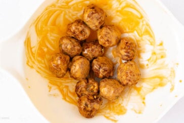 A white plate containing twelve round, evenly coated meatballs in a glossy orange sauce, with some sauce smeared around the plate.