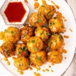 A plate of meatballs topped with melted cheese and garnished with chopped herbs. A small dish of red sauce is placed beside the meatballs, and drizzles of sauce decorate the plate.