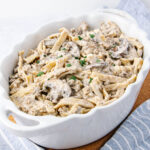 A white casserole dish filled with creamy beef and mushroom pasta, topped with chopped parsley. The dish rests on a light blue and white striped cloth.
