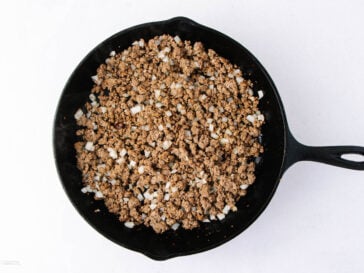 Ground beef with diced onions is cooking in a black skillet on a white background.