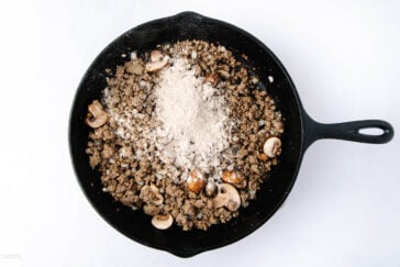 A black skillet containing cooked ground beef, sliced mushrooms, and a mound of flour on top—perfect for starting a classic beef stroganoff with ground beef—set against a white background.