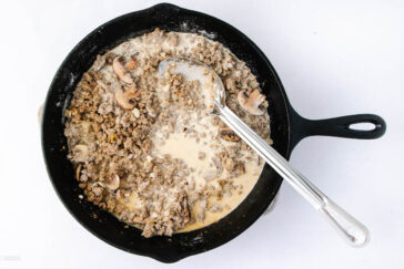 A black skillet on a white surface contains cooked ground beef with mushrooms in a creamy sauce. A metal spoon rests in the pan, partially submerged in the mixture.