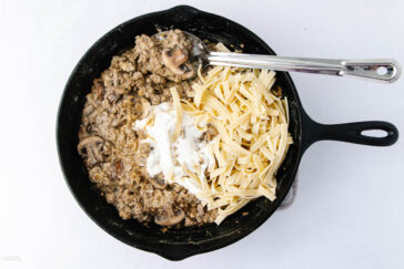A black skillet contains a mixture of cooked ground meat and sliced mushrooms on one side, with uncooked pasta and a dollop of cream or sauce on the other. A metal spoon rests in the skillet.