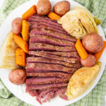 A plate of sliced corned beef surrounded by boiled red potatoes, carrots, and wedges of cabbage, all arranged on a white plate. The meal is set on a green checkered cloth.