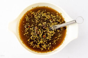 A white bowl filled with a dark liquid marinade, containing herbs, spices, and peppercorns. A small metal whisk with a loop handle rests inside the bowl. The background is a plain white surface.